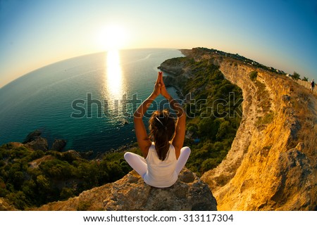 Young slim Yoga woman in zen meditating in warrior pose relaxing outside on top of mountains and sea on sunrise or sunset. Asian fitness model doing yoga on blue sky and blue sea background.