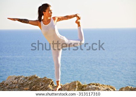 Young slim Yoga woman in zen meditating in warrior pose relaxing outside on top of mountains and sea on sunrise or sunset. Asian fitness model doing yoga on blue sky and blue sea background.