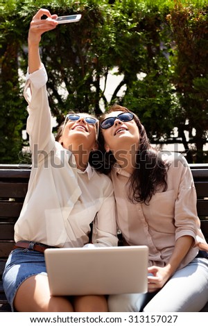 Two beautiful girls sitting on a bench in the street and make selfie-photo. Two students with a laptop resting on skayke in the city, laughing and doing selfie.