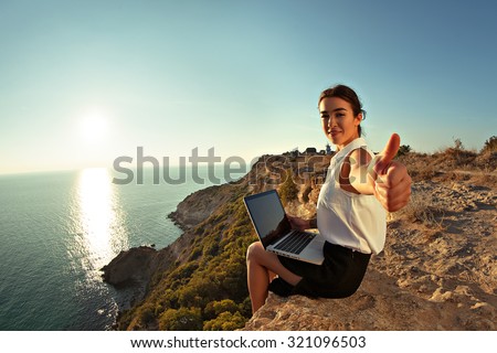 Young business woman working at the computer on the beach on the rock face and shows Like. Young girl working at a laptop at sunset or sunrise on the top of mountain to the sea. Downshifting concept.