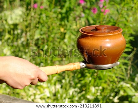 Oven fork and clay pot in woman hand against green grass background.