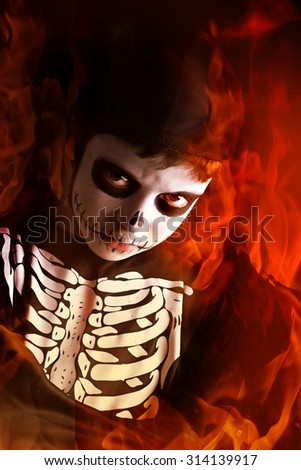 Boy with face-paint and skeleton Halloween costume isolated in a dark background with the flames of hell