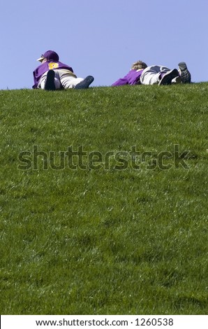 Two baseball players lie on the top of a grassy hill watching a baseball game.