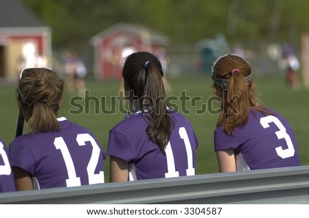 Three girls sit on the bench at a lacrosse game.