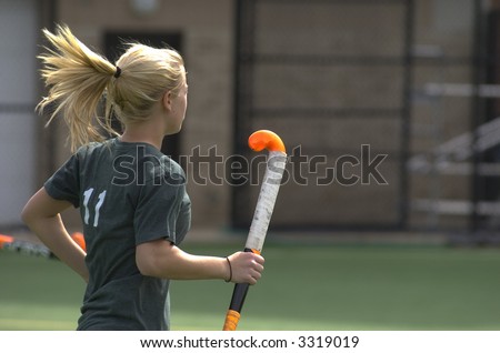 A blond, teenage girl runs across the field holding a bright orange field hockey stick.