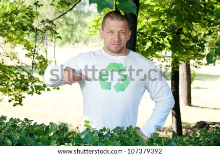 An adult man standing in green park and pointing at recycle logo on his shirt