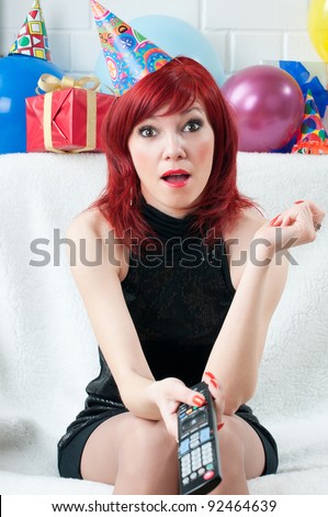 Surprised red-haired party woman sitting before TV with remote control