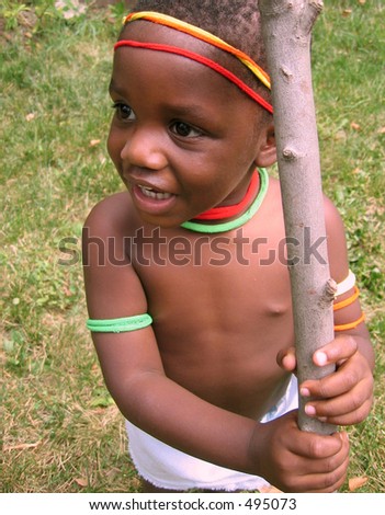 Small African boy wearing colored decorations holding a stick.