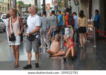 HOLLYWOOD, CA - AUGUST 3, 2014: Tourists stroll the Walk of Fame in Hollywood as two girls sit on the sidewalk, arms raised holding cell phones, taking photos of themselves near a celebrity star.