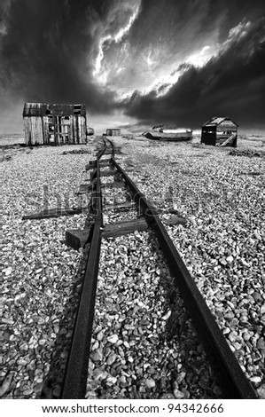 narrow gauge rails running through abandoned sheds, boats and equipment on a shingle beach in black and white with a moody sky beyond