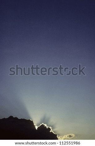 Sun silhouettes a dark cloud with bright rays in blue sky
