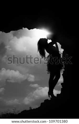 Silhouette of the slim lady standing in the dark cave against the cloudy sky and sun