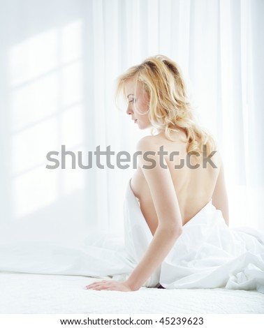 Young blond lady in a bedroom with window shadows on the wall. Rear view