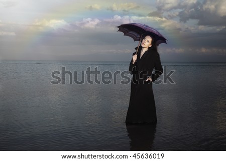 Sensual lady in the black coat with umbrella standing in the water after the rain. Rainbow on a background