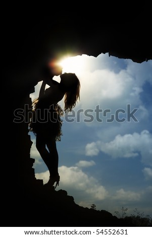 Silhouette of the slim lady standing in the dark cave against the cloudy sky and sun