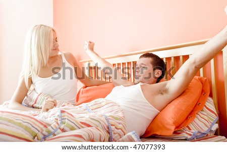Man stretches while reclining in bed as woman sits and smiles at him. Horizontal format.