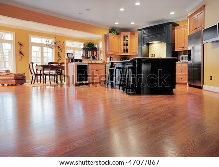 Home interior shows a large expanse of wood flooring in the foreground and a kitchen and dining room in the background. Horizontal format.