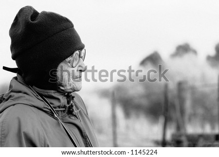 An elderly woman, dressed in winter clothes and hat, stares off into the distance.