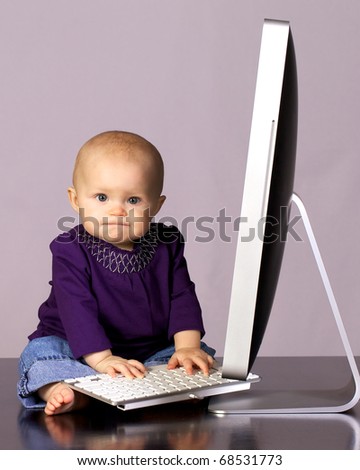 Infant baby girl sitting up looking playing with a desktop computer screen and keyboard.