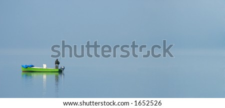 A fisherman stands alone in his boat on a misty lake, shortly after dawn. Space for text.