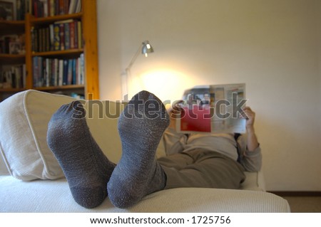 A man lies on the couch with his feet up, reading the paper. Focus on the slightly worn socks. Space for text on the wall, top right.