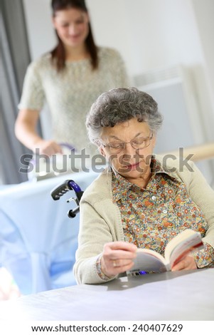 Elderly woman reading book while home helper irons laundry