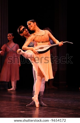 female classic ballet dancer dressed white dancing holding mandolin held by male classic ballet dancer dressed brown lit by stage lights