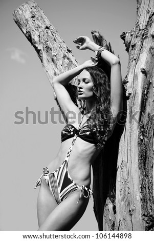 Fashionable woman in bikini leans one\'s elbows on a tree. Outdoor black-and-white portrait.