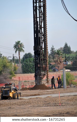 A power auger to drill deep holes is supported by a crane on a construction job site