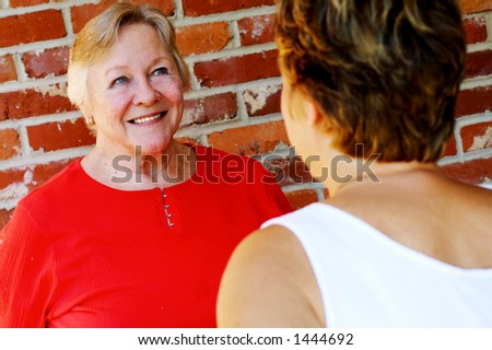 Two women smiling and standing in front of a wall. One woman is saying something funny. Shallow depth of field, focus on woman on left. Woman on right slightly out of focus.