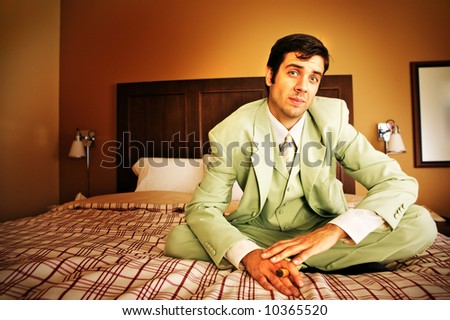 A businessman in a green suit relaxing on his hotel room bed about to enjoy a cigar.