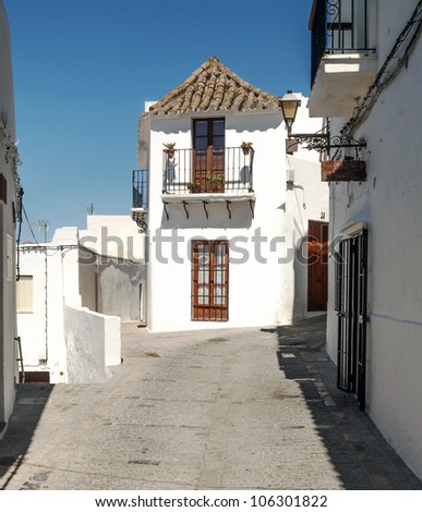 Close street decorated with white houses and bars in their windows. In the background are two paths. It is Situated in a village in Spain