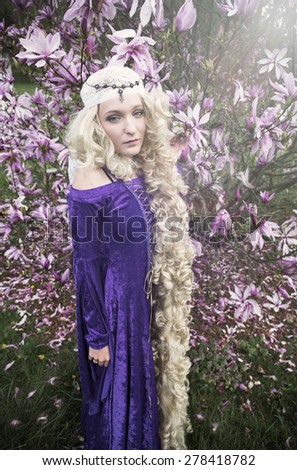 Young woman dress as Rapunzel wearing purple velvet gown standing in front of magnolia tree with long curly blond hair.