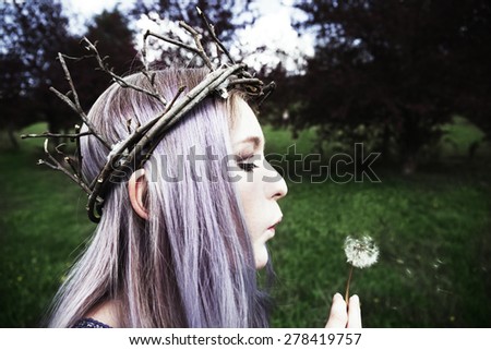 Young woman with purple hair blowing on dandelion and wearing twig crown
