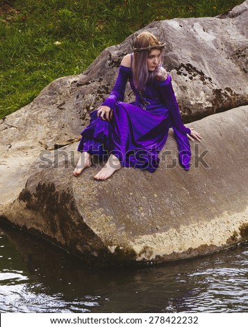 Young woman wearing velvet purple gown and twig crown sitting on rocks by a river