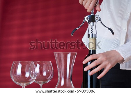 Women opening red wine by corkscrew. Next to her stay two wineglasses and carafe. Red background behind her with sun beams and shades.