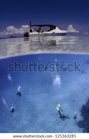 Scuba divers swimming underwater with white diving boat on the ocean surface. Horizontal split over/under color image.