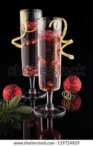 Closeup of glasses of pomegranate champagne cocktail, garnished with lemon twist, and Christmas ornaments on black background.