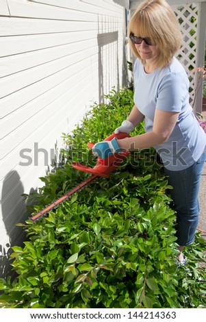 Woman trimming bushes in her backyard using an electrical hedge trimmer.