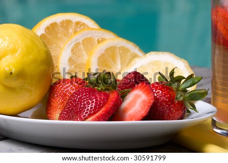 Glass of home made iced cold pink strawberry lemonade and plate full of fresh lemons, strawberries on hot summer on edge of swimming pool.