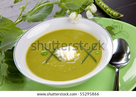 Bowl of pea soup garnished with chives and cream. Sprig of flowering pea and cracked pea pods.
