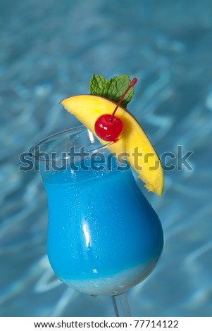 Glass of Blue Hawaiian cocktail on swimming pool side garnished with mango wedge and maraschino cherry