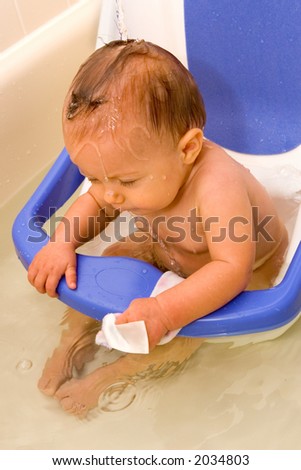 Baby in bathtub safety seat, holding on while her hair is rinsed with fresh water after washing her hair. Captured with high speed strobe to freeze the motion of the poured water and drops.