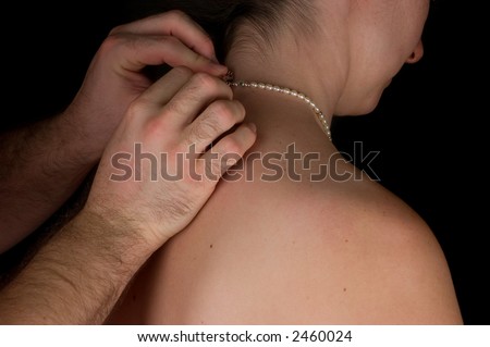 Man helps a young woman put on her pearl necklace in a darkened room.