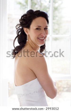 Beautiful brunette bride in white wedding dress stands by a bright sunlit window giving a lovely high key effect.