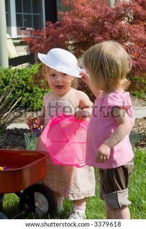 One year old baby girls playing in the yard. Girl in dress is smiling as her friend places a white hat on her head. Image about themes of friendship and childhood.