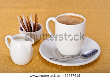 Mug of coffee with saucer, teaspoon, milk jug and sugar sachets on a golden table cloth