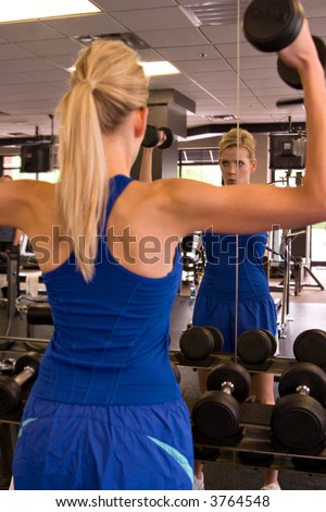 Beautiful blond woman lifting weights while looking in a mirror in a fitness center. Reflection in mirror is in focus - foreground image is out of focus.