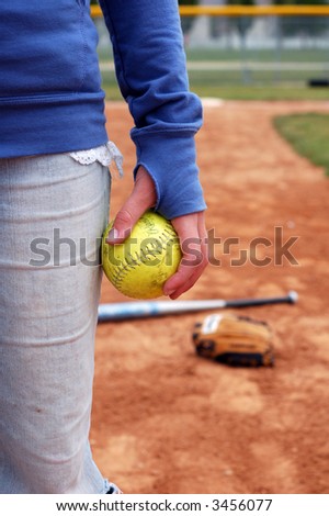 A young woman holds a softball.  Mitt and bat are out of focus on the infield.