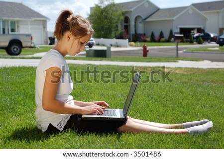 A young woman types on her computer in the grass.